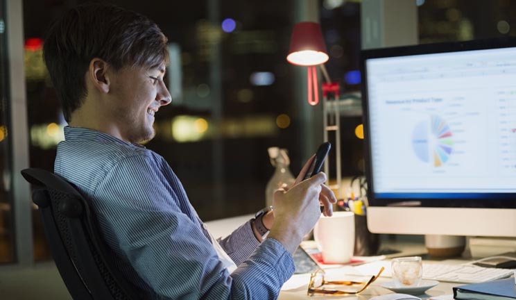 Man sitting at computer screen looking at his phone 